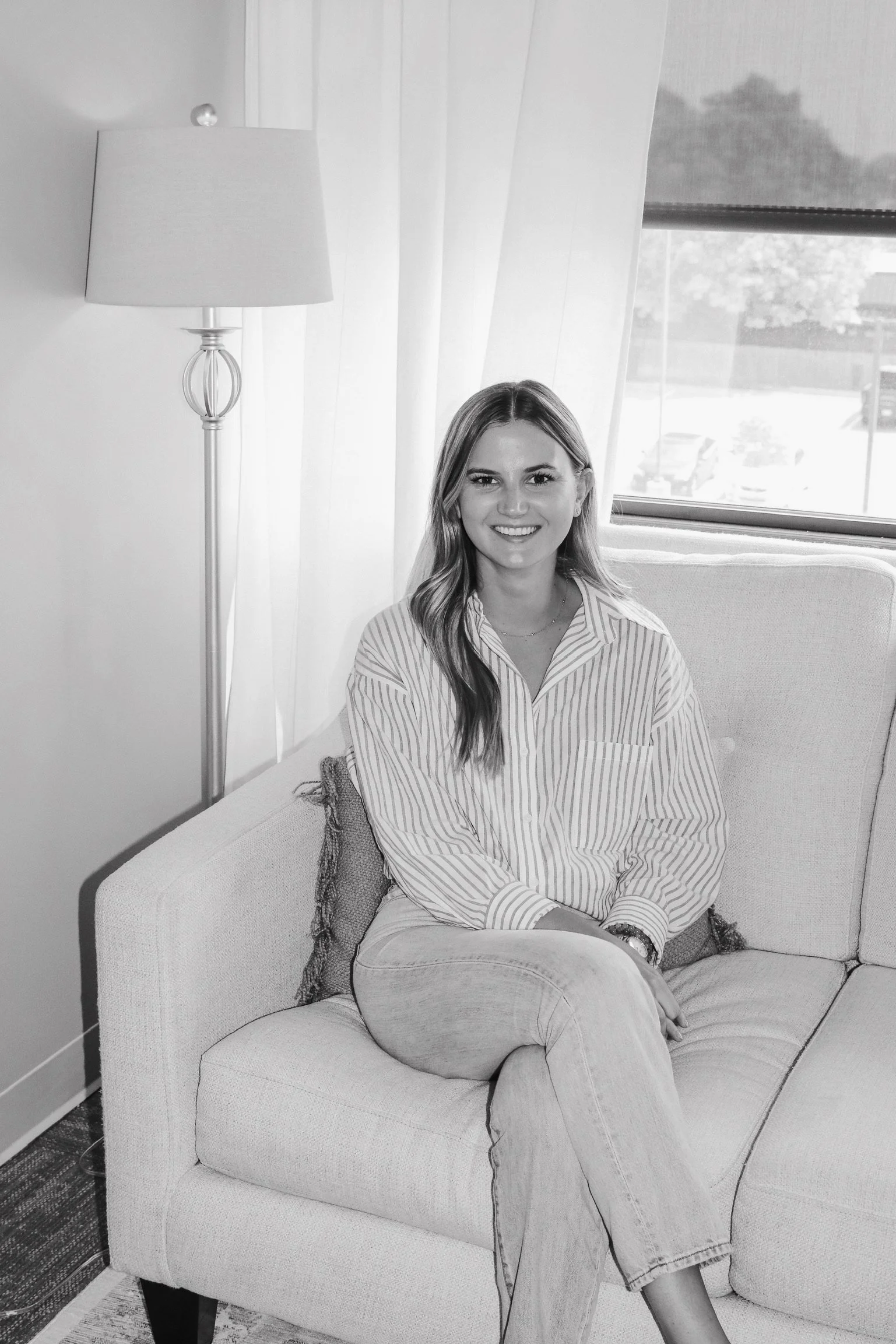 A therapist in Lenexa Kansas  sitting on a couch in a well-lit room, wearing a striped shirt and light-colored jeans.