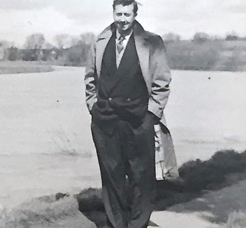 Black and white photo of a man standing outdoors near a body of water, wearing a coat, vest, and trousers, with trees and an open landscape in the background.