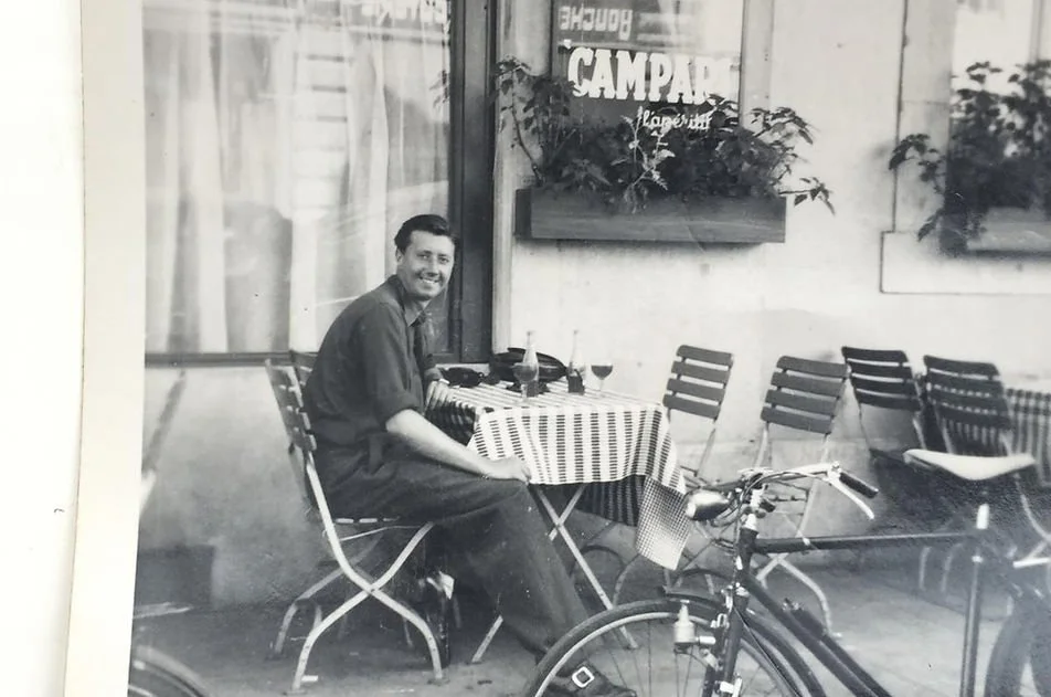 A man sitting at an outdoor table with a checkered tablecloth, smiling at the camera. There are several empty chairs around the table, a bicycle in the foreground, and potted plants on the wall behind him. The setting appears to be a restaurant or cafe with large windows and a sign with partially visible text.