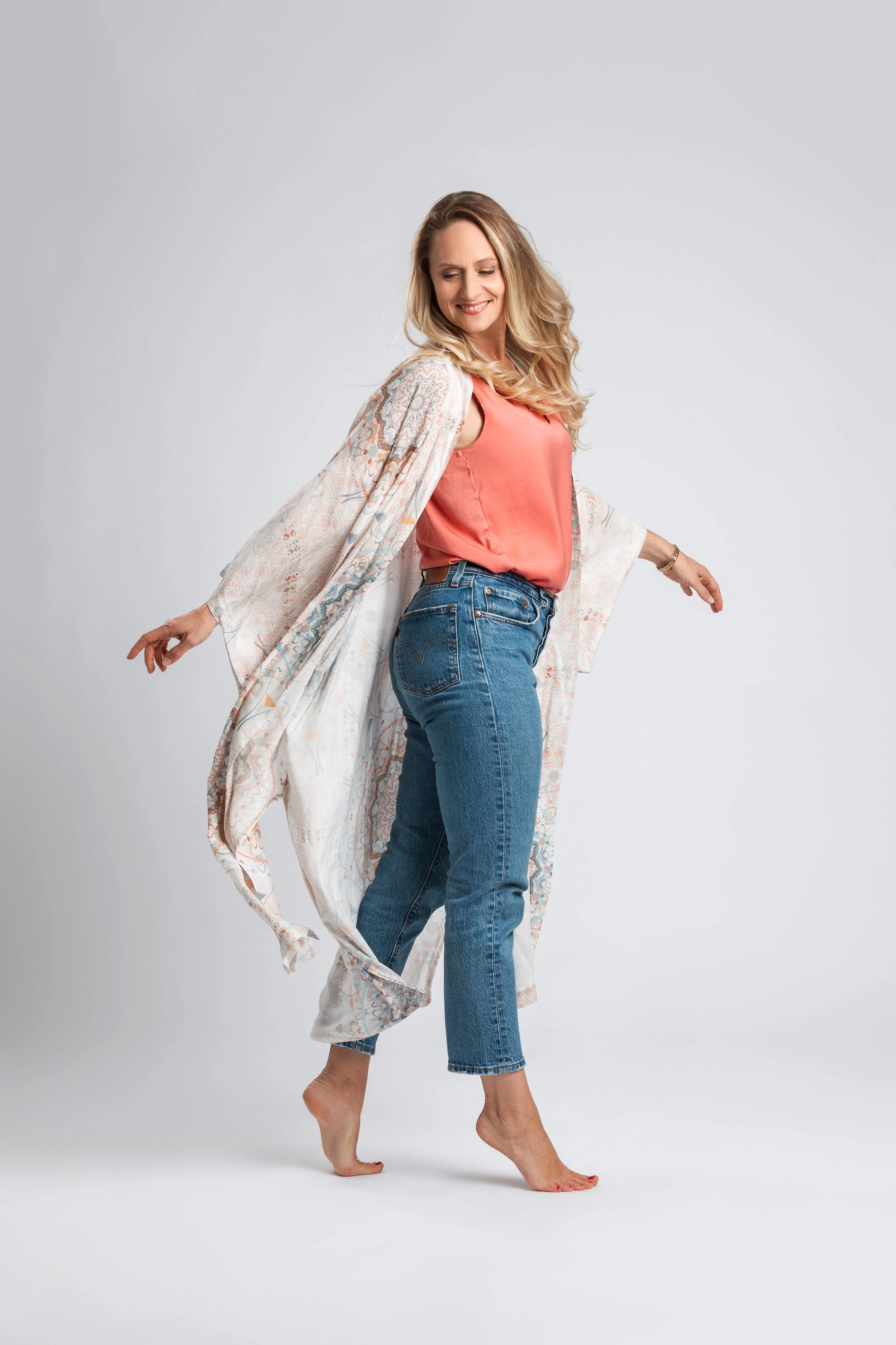 Eine lächelnde Frau mit langen blonden Haaren, die in Jeans und einem rosa Top steht, während sie barfuß auf einem weißen Hintergrund tanzt oder posiert.