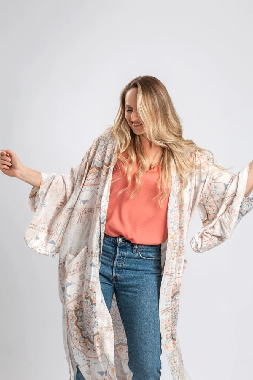 Frau mit langer, blond gewellter Frisur trägt eine peachfarbene Bluse, dunkle Jeans und einen langes, gemustertes Kaftan, tanzt vor grauem Hintergrund.