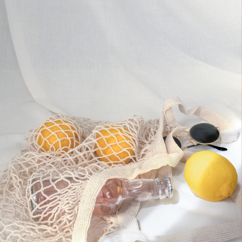 A mesh shopping bag holding oranges, with a lemon and a clear plastic bottle nearby, placed on a white surface.