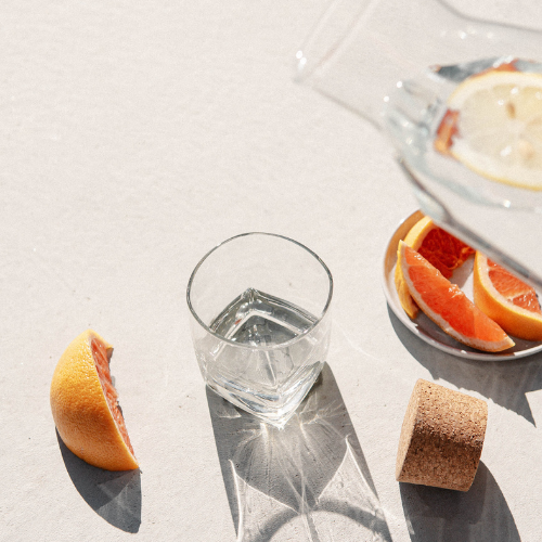 Glass water, grapefruit slices on a plate, a cork, and a glass jug with lemon slices on a light surface.