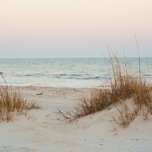 Beach with white sand, some grass on dunes, calm ocean waves, and a pastel-colored sky.