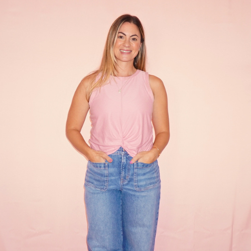 A young woman with long blonde hair wearing a pink sleeveless top and blue jeans standing against a light pink background, smiling with hands in her pockets.