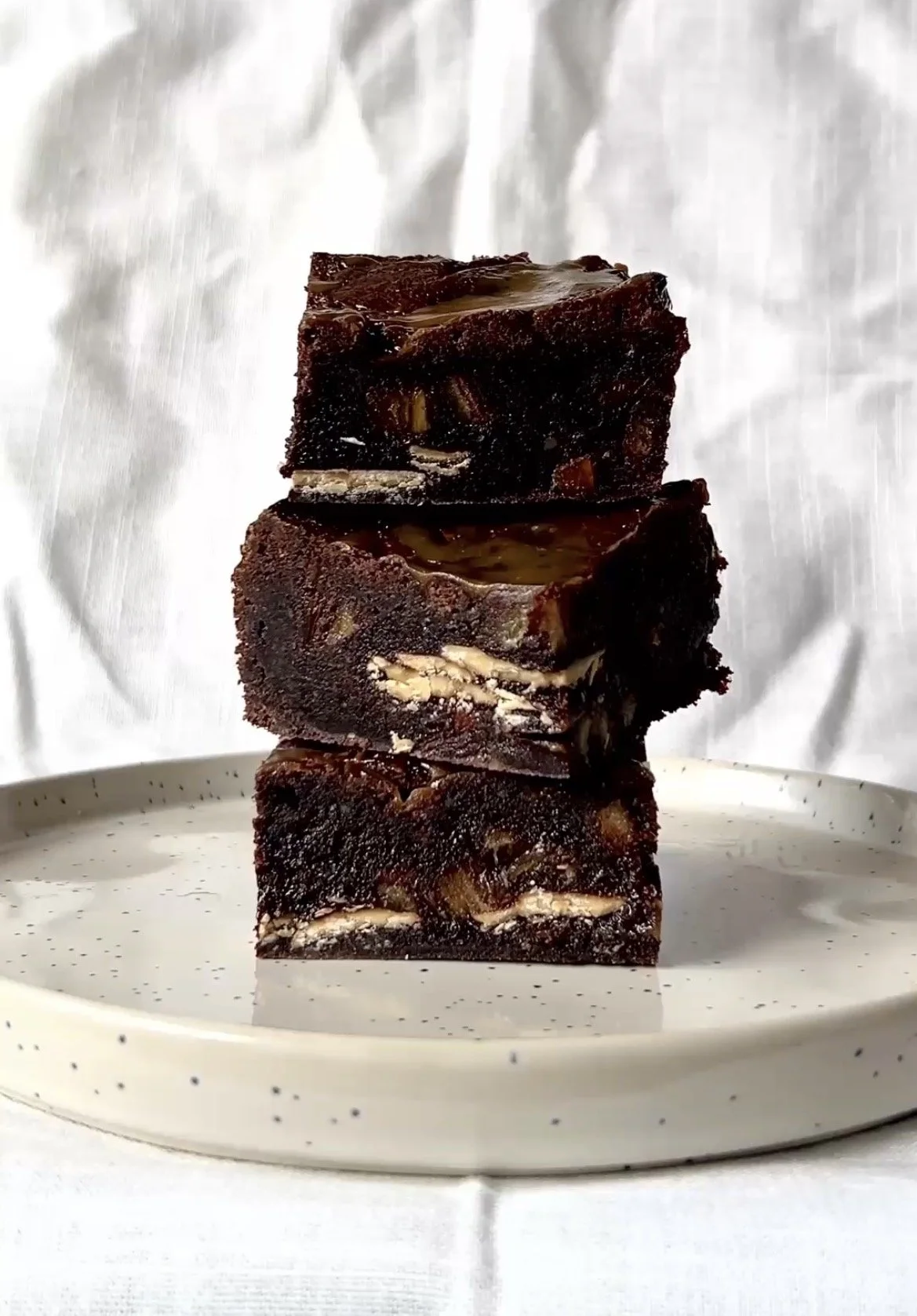Three stacked pieces of chocolate brownie with nuts on a white speckled plate, against a white background.