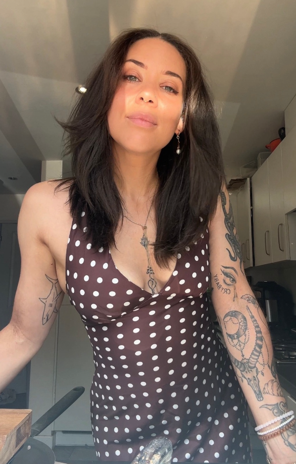 A woman with dark hair, light skin, and tattoos on her arms, wearing a black and white polka dot sleeveless dress, standing in a kitchen.