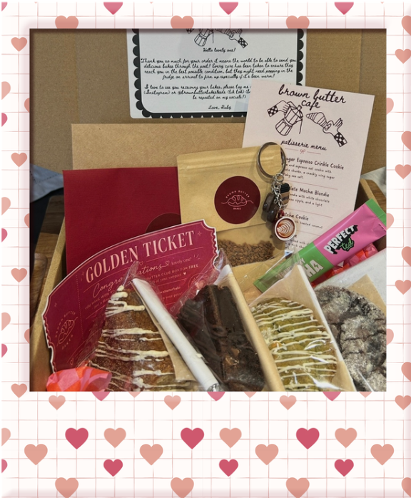 A gift box with assorted baked goods including donuts and cookies, a red Golden Ticket card, a small bag of granola or tea, a white pen, a pink gum pack, and a keychain with a small charm, placed on a heart-patterned background.