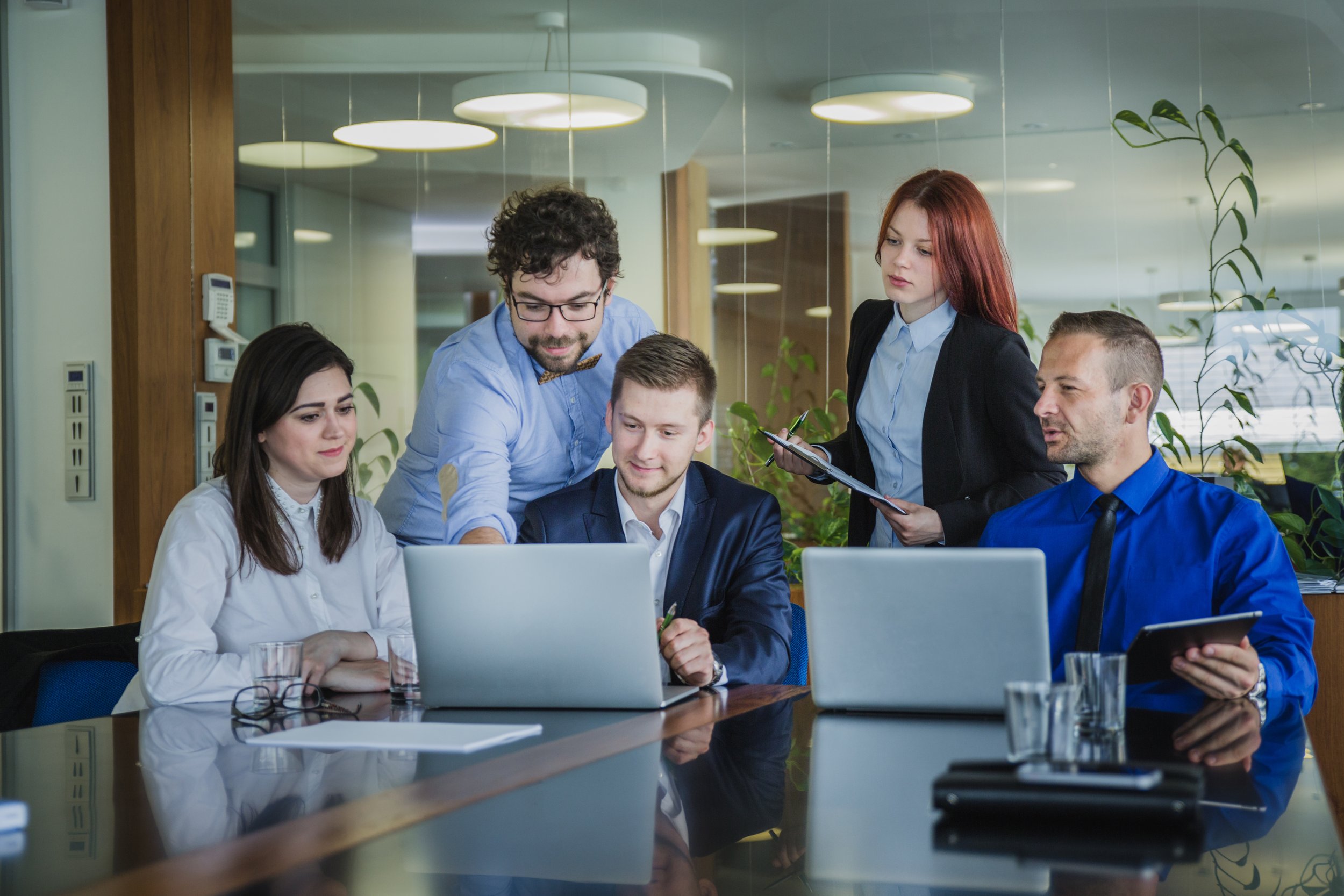 A group of five diverse business professionals gathered around a table in a modern office, working together on a laptop and discussing a project.