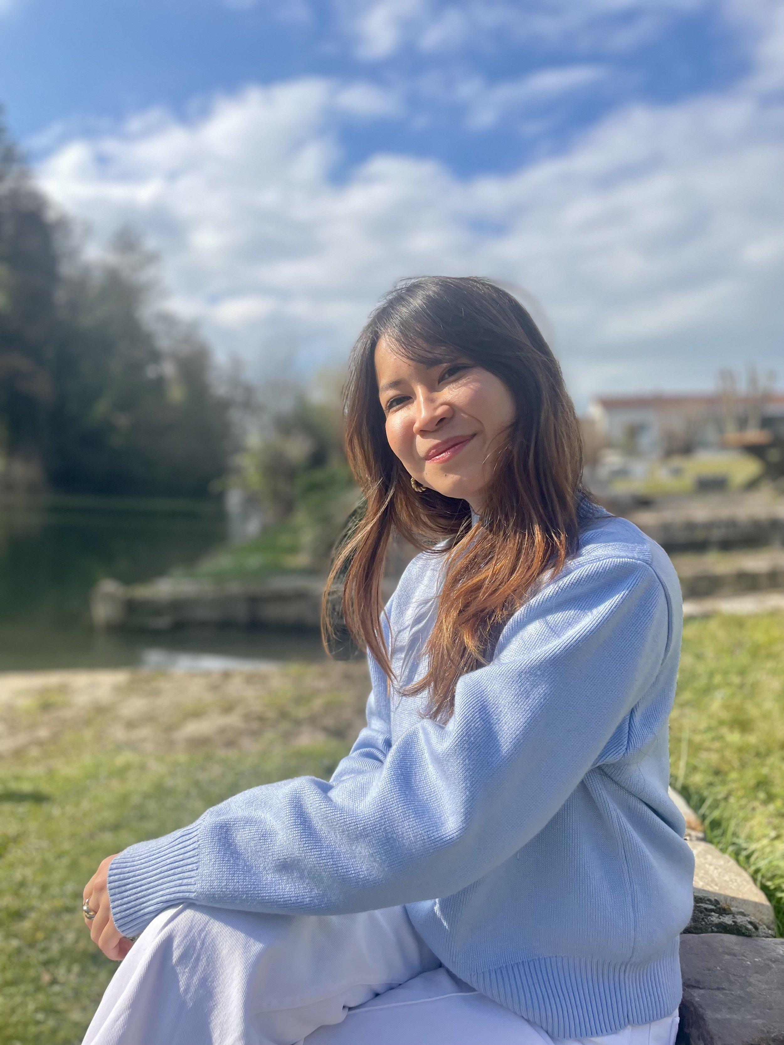 Jeune femme avec des cheveux bruns ondulés, assise sur un rocher dans un parc, sous un ciel bleu avec quelques nuages, portant un sweat bleu clair et des vêtements blancs, souriant doucement.