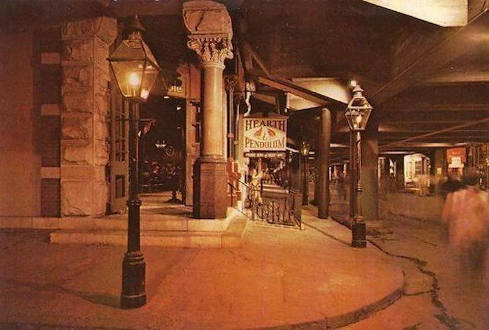 Under an overpass, a corner of a brick building with a sign for 'Hearth & Pendulum' is visible. There are vintage-style street lamps and a person walking by.