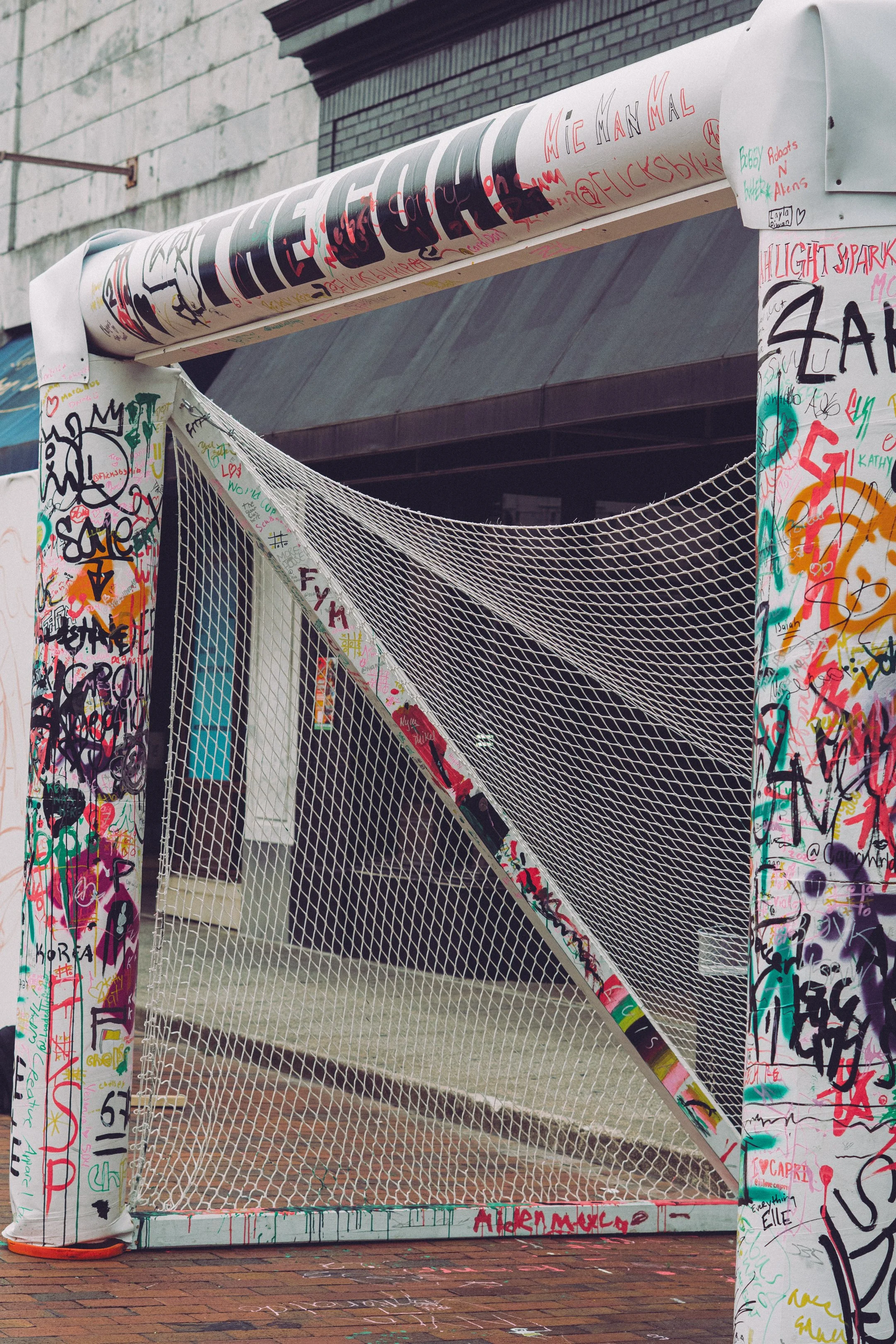 A sports goalpost covered in colorful graffiti, with a baseball net hanging from it, situated on a brick sidewalk with a concrete wall in the background.