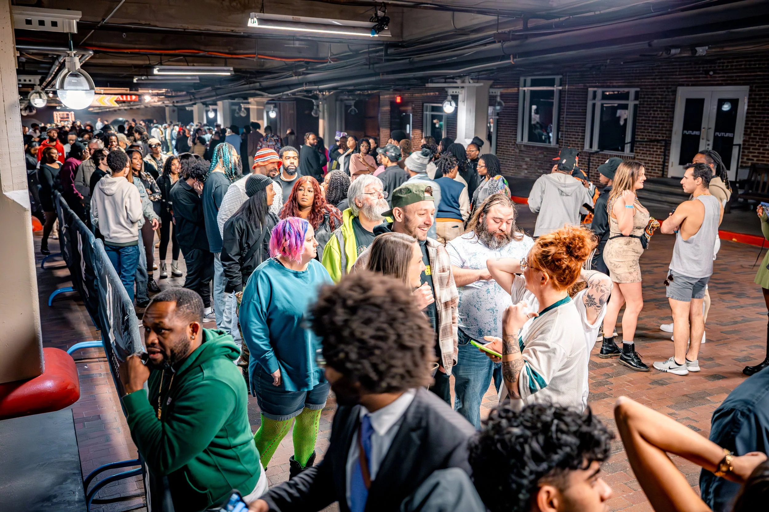 Crowd of diverse people standing and walking in an indoor space with brick walls and ceiling pipes, some engaging in conversations, with a few sitting or leaning on benches.