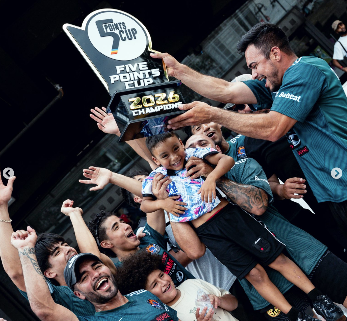 A group of people celebrating with a child being lifted and holding a trophy that reads 'Points Cup' and '2026 Champions.' The scene appears to be a sports victory celebration, with smiling faces and raised hands.