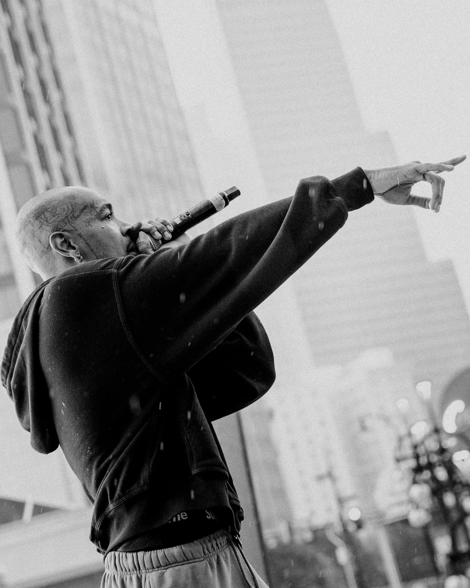 A person with a shaved head and tattoos near the eye, holding a microphone close to their mouth, pointing forward with their other arm extended, wearing a dark jacket and light-colored sweatpants, in an outdoor urban setting with buildings and blurred lights in the background.