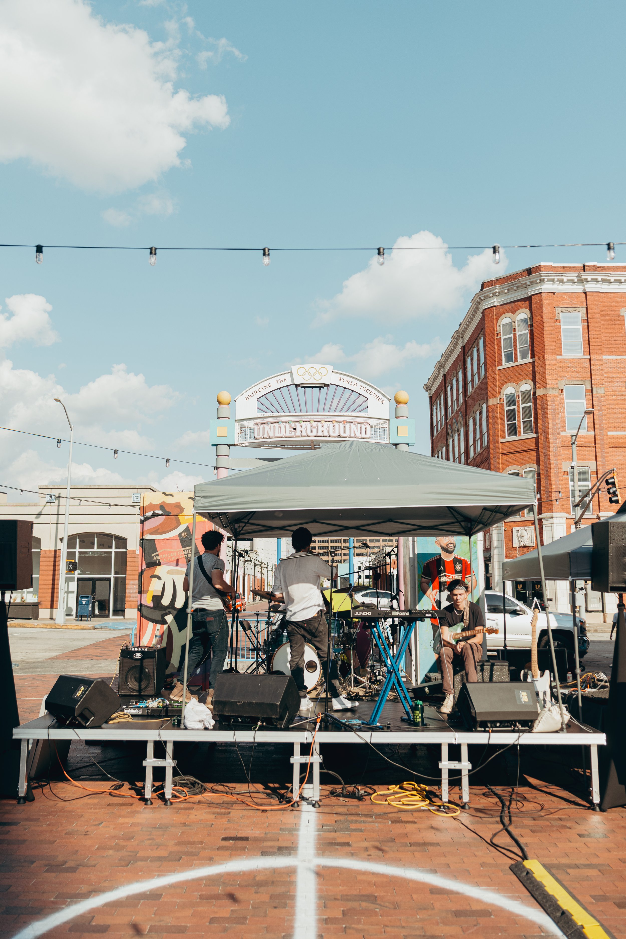 Band performing live music on a stage outdoor in an urban setting with brick buildings, hanging string lights, and a blue sky with clouds.