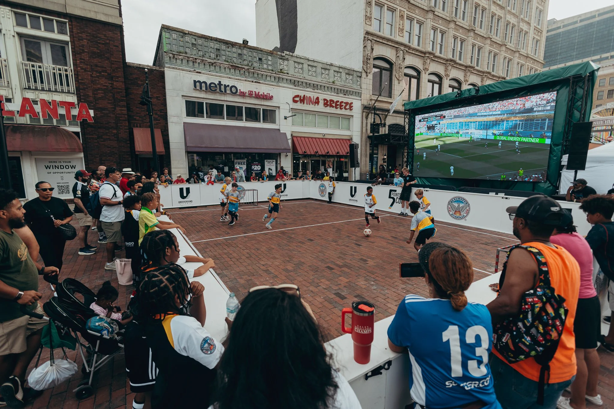 People watching a soccer game projected on a large outdoor screen at a street event in Downtown Atlanta - Underground Atlanta., with children playing soccer on a small court nearby.