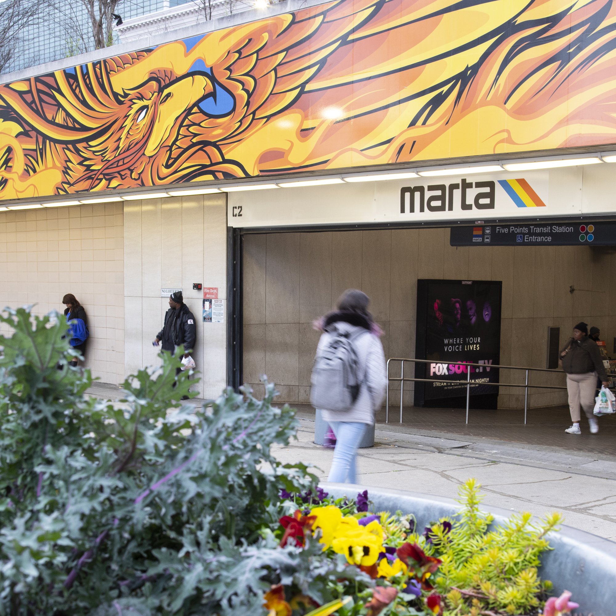 Colorful mural of a phoenix on the exterior wall of a building at Five Points Transit Station, with people walking past and flowers in the foreground.