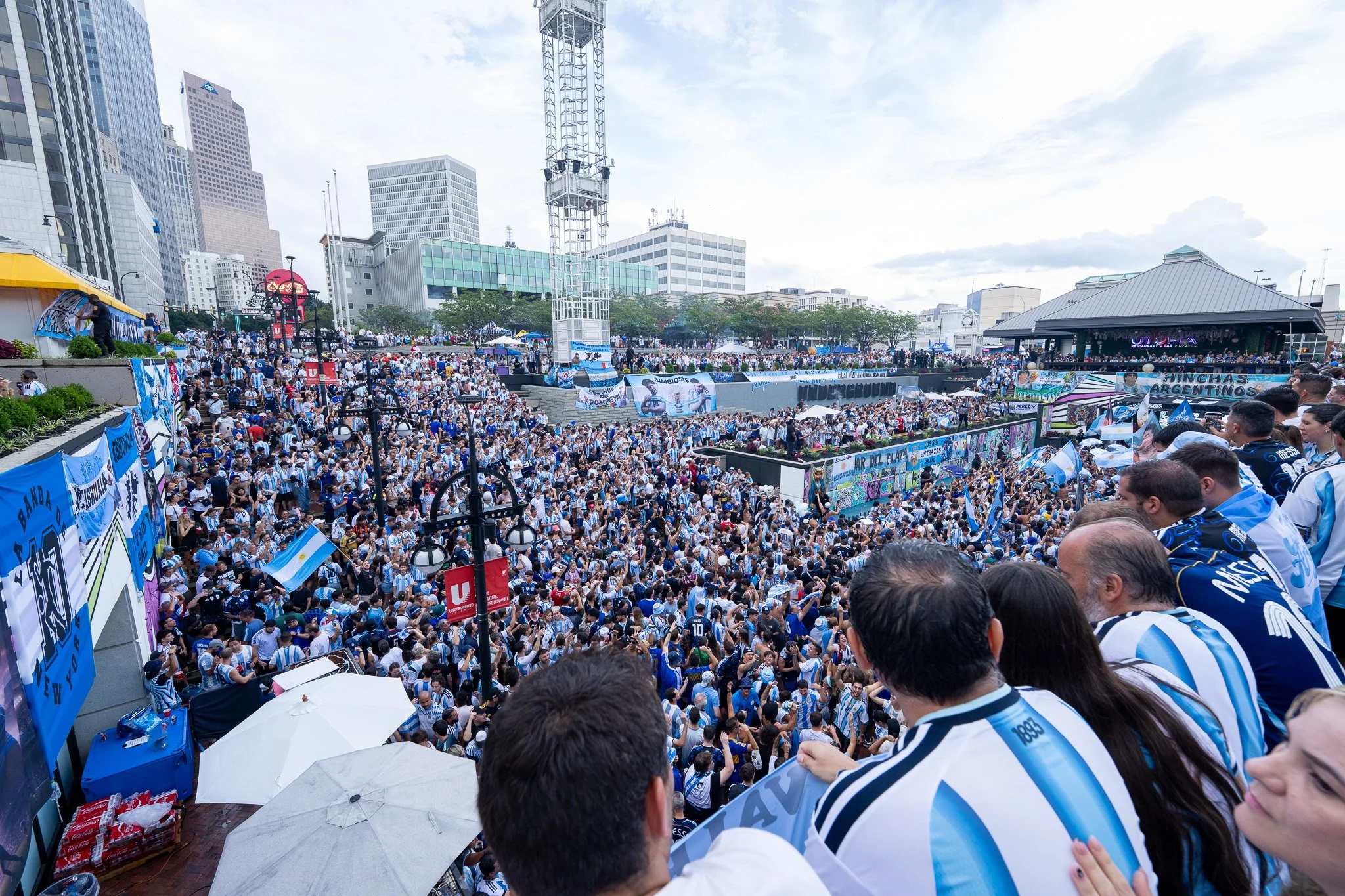 Crowd of people gathered at a sports event in an urban area, many wearing blue and white jerseys, with banners and flags, in front of tall buildings and a stage with a band.
