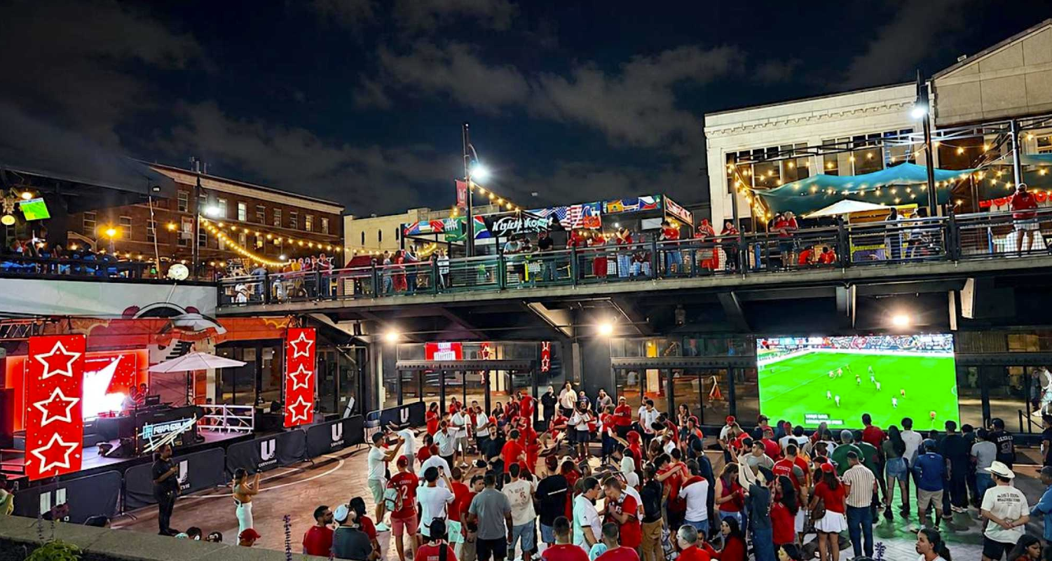 Night scene of a lively outdoor sports bar with many people gathered to watch a soccer game on a large screen, decorated with red, white, and blue flags and string lights.