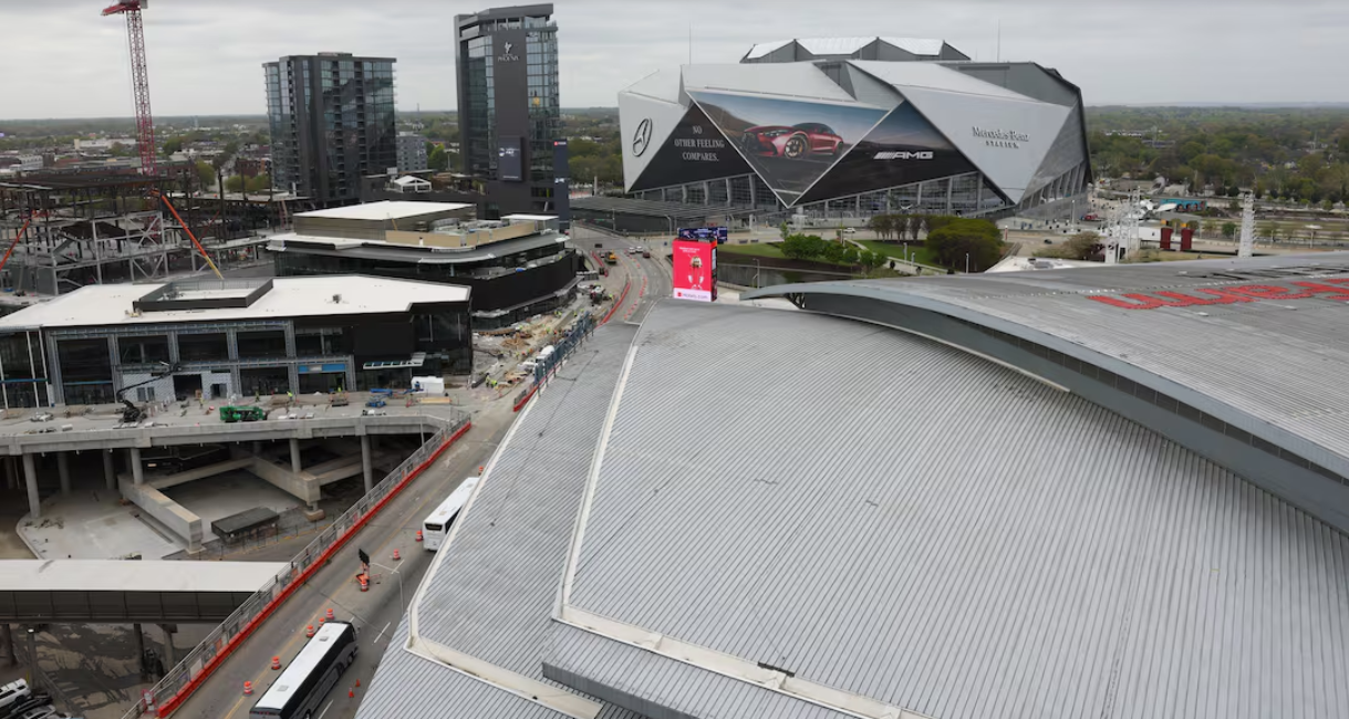 An aerial view of a cityscape with tall buildings, a large stadium with a curved roof, and a construction site with a crane.