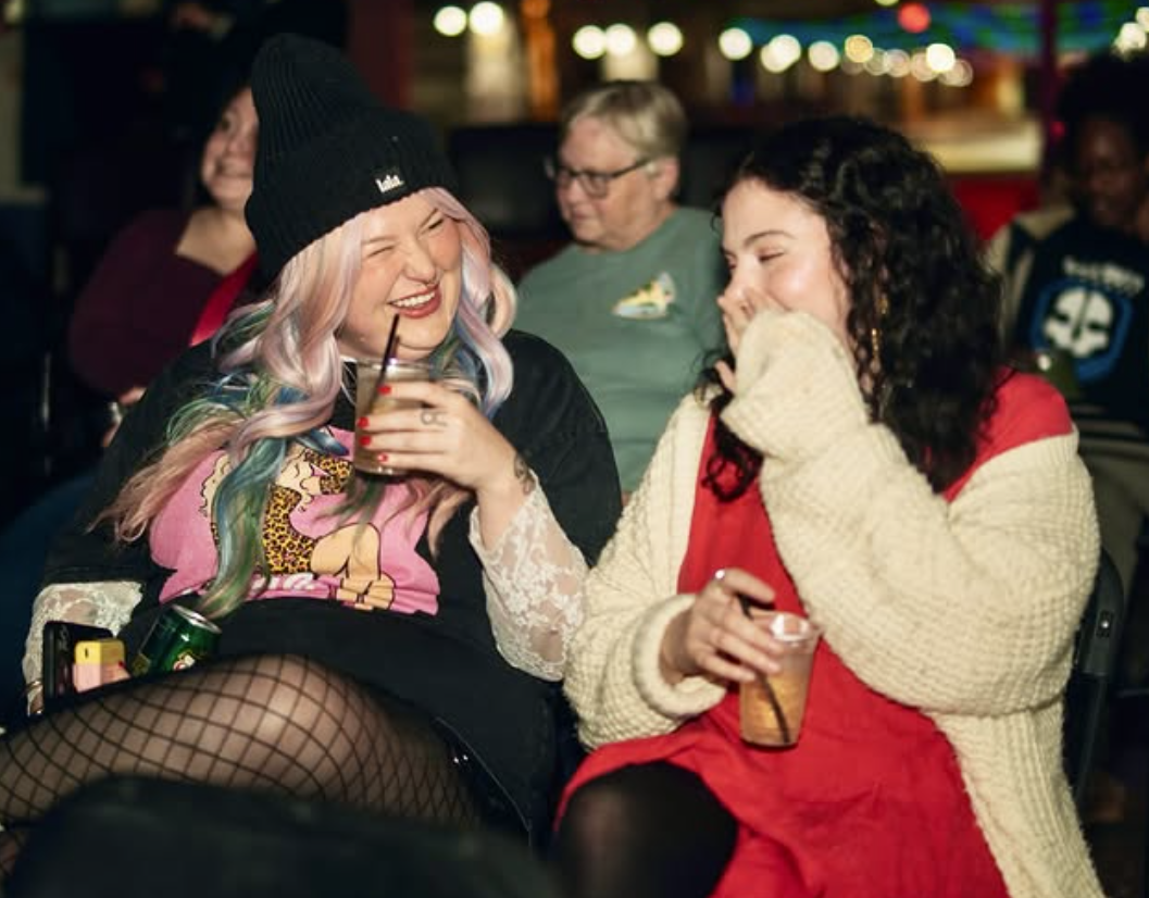Two women laughing and enjoying drinks at a social gathering with colorful lights in the background.