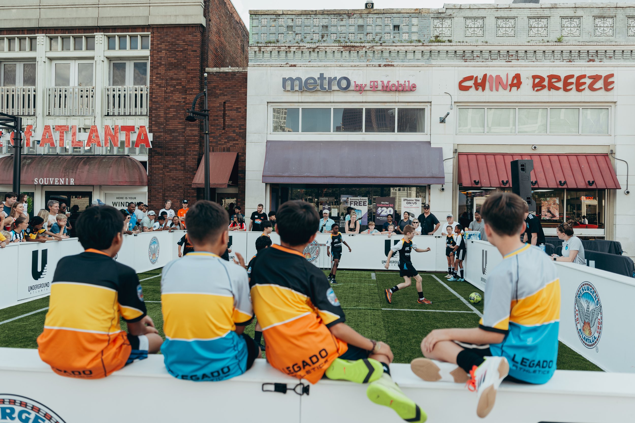 Children playing soccer on a field during an outdoor event, with spectators and buildings in the background.