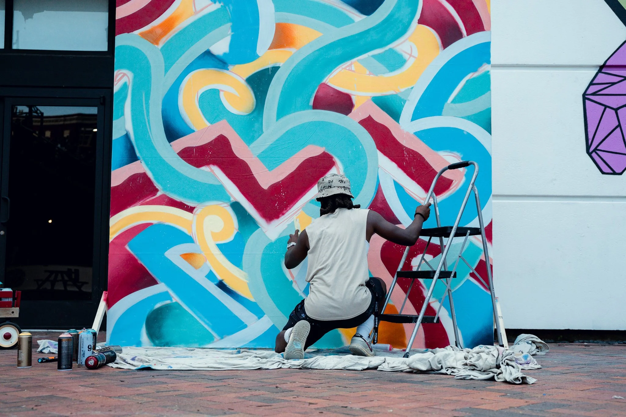 An artist creating a colorful abstract mural on a large outdoor wall, with spray paint cans, a step ladder, and painting supplies on the ground around them.