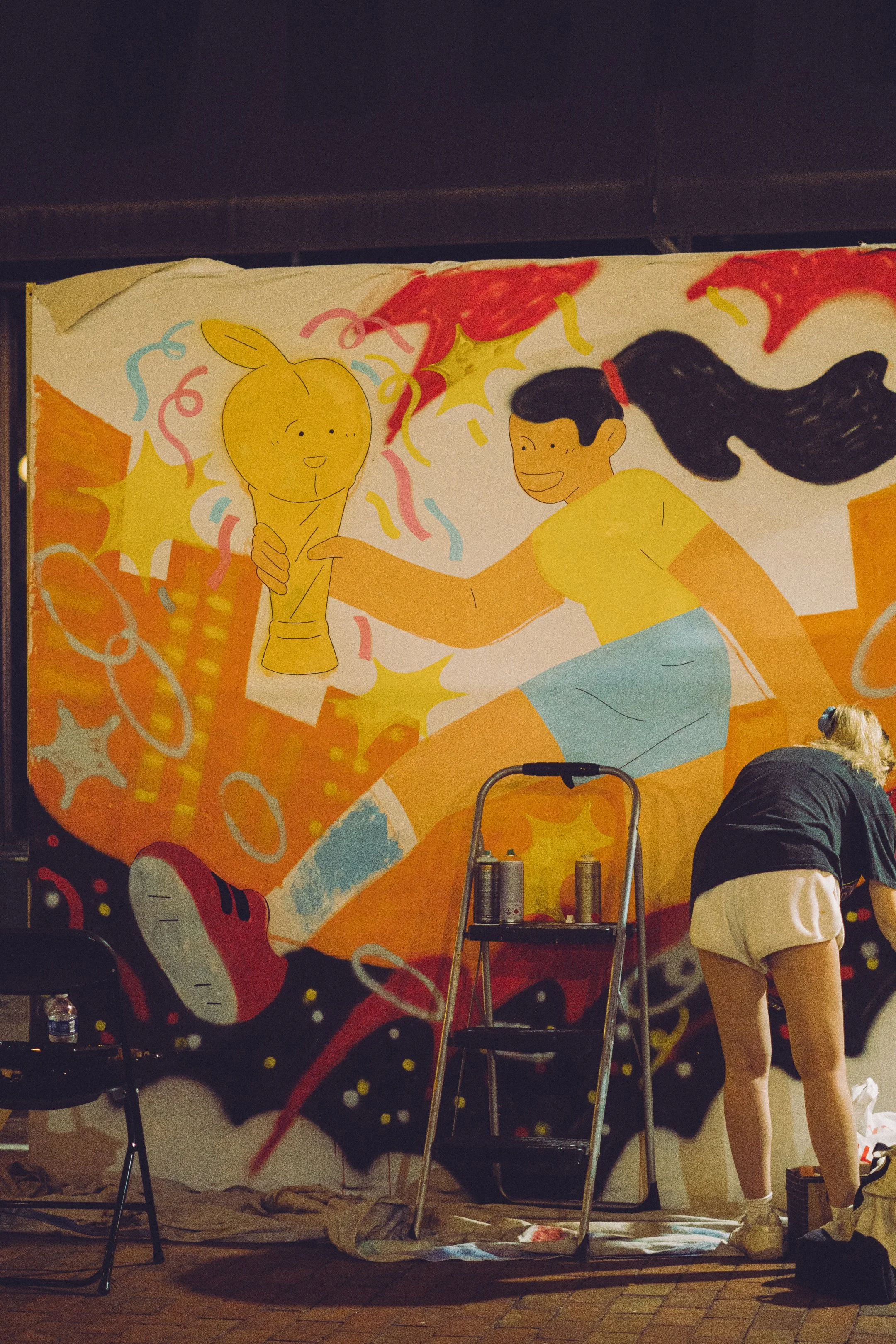 A person in beige shorts and a black shirt creating a colorful mural on a wall, with art supplies and a step ladder nearby, in a dimly lit indoor space.
