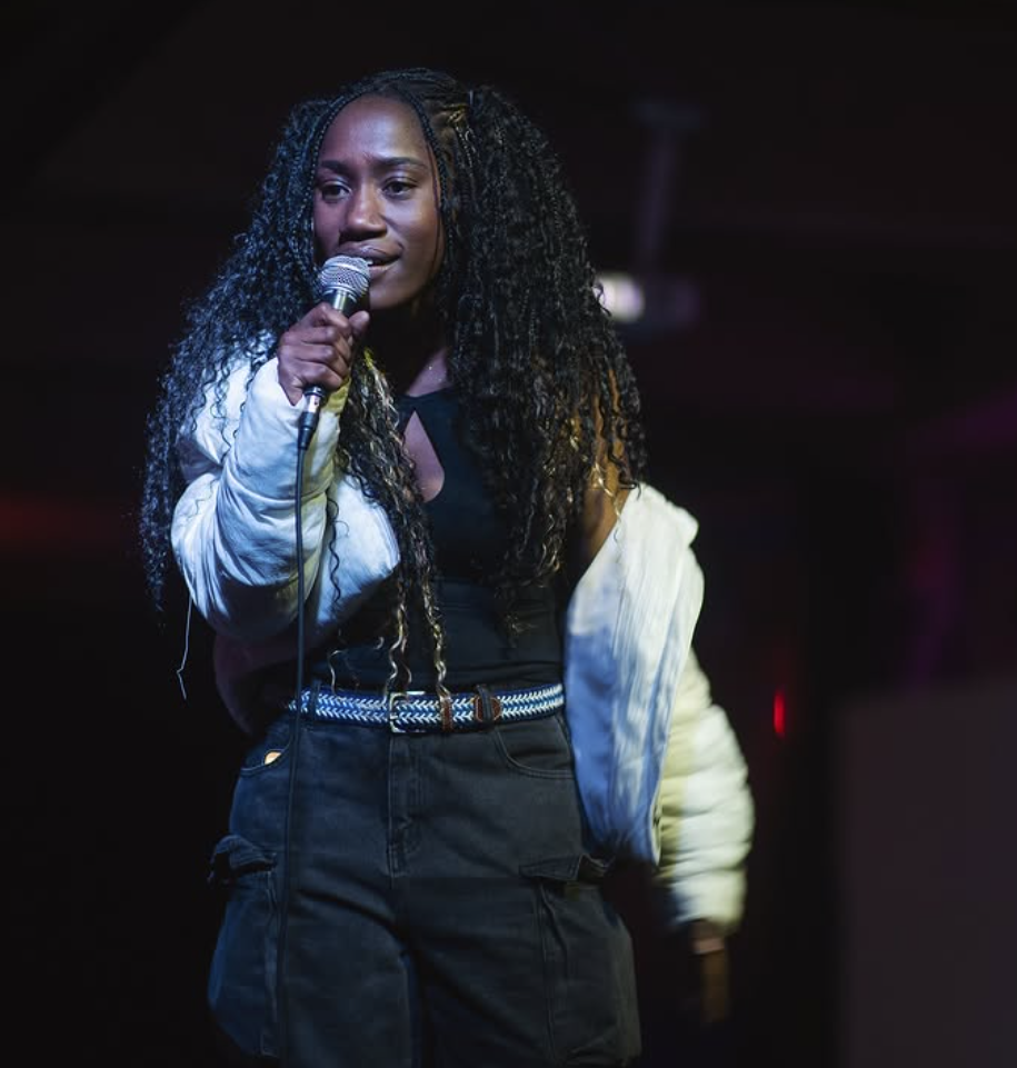 A woman with long curly hair holding a microphone on stage, wearing a black top, black pants, and a light jacket.
