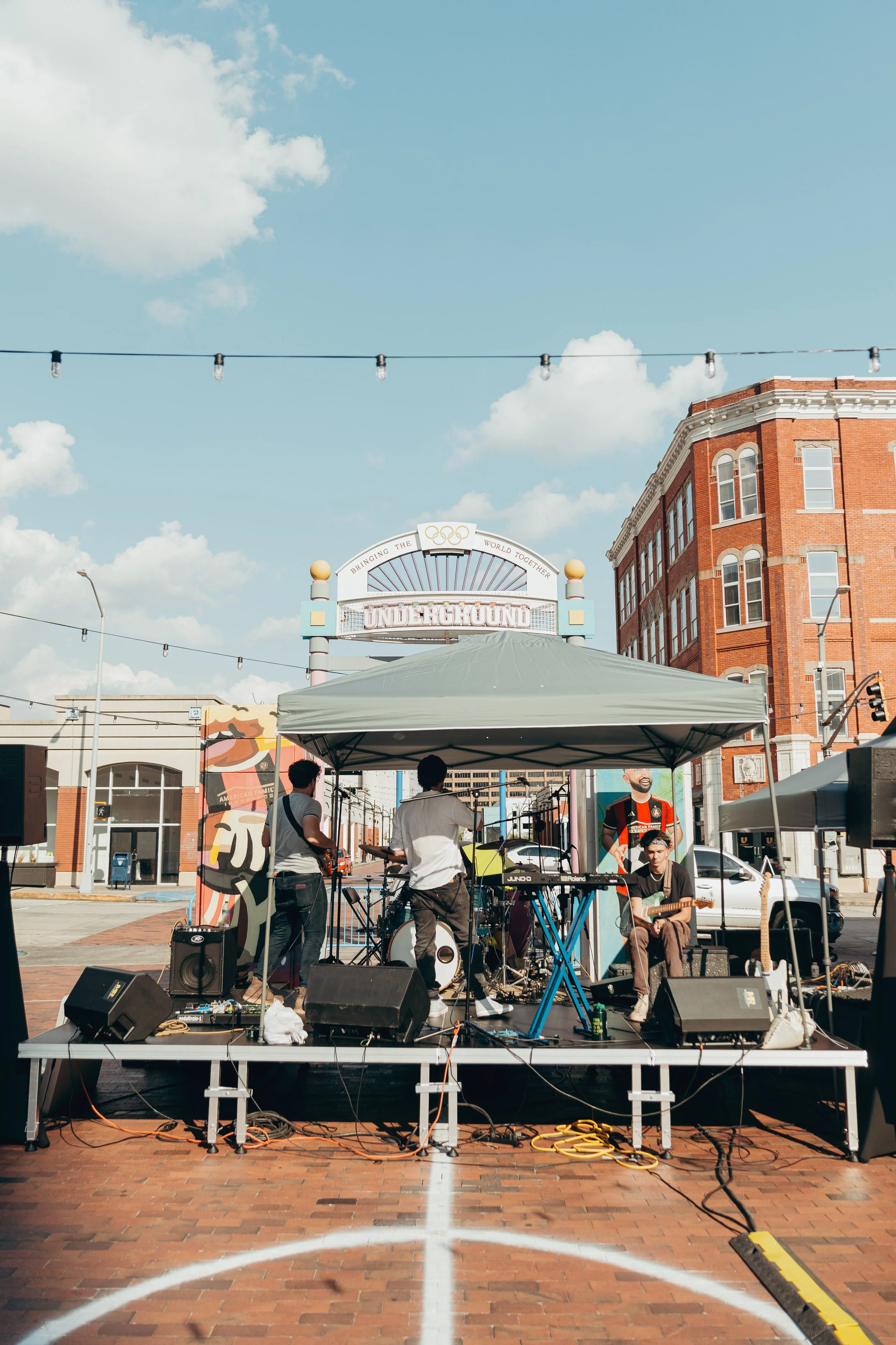 A live outdoor musical performance on a stage set up in a city square. Four band members are playing instruments; one on keyboard, one on guitar, and others are either singing or playing percussion. The stage has a canopy, and the audience is not visible. In the background, there's a building with brick walls, the Olympic rings logo and a sign that says 'Underground'. The sky is partly cloudy with string lights hanging overhead.