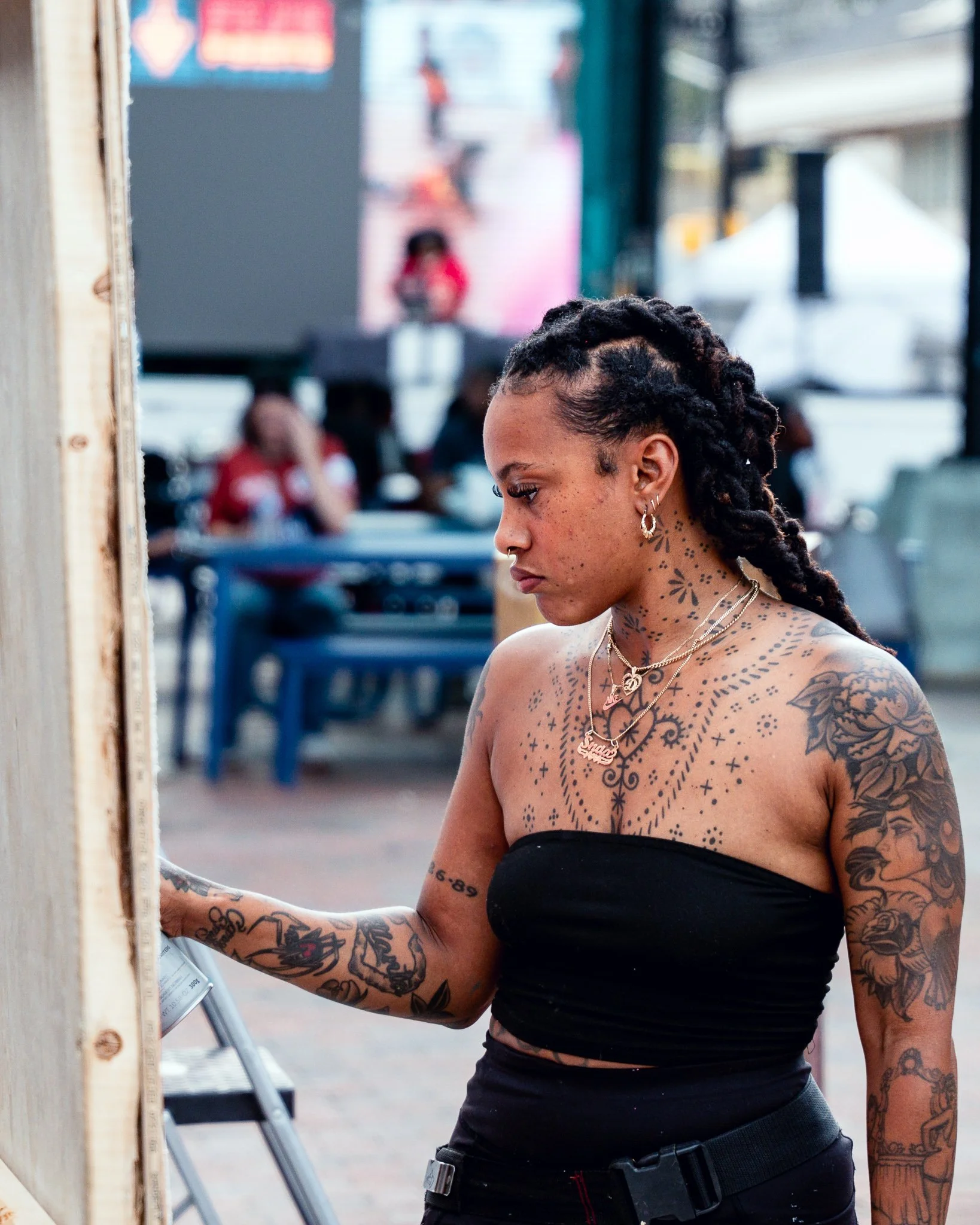 A woman with tattoos on her arms and chest, wearing a black strapless top and jewelry, is looking down and to the right. She has braided hair and is standing outdoors near a wooden structure, with people and a screen in the blurred background.