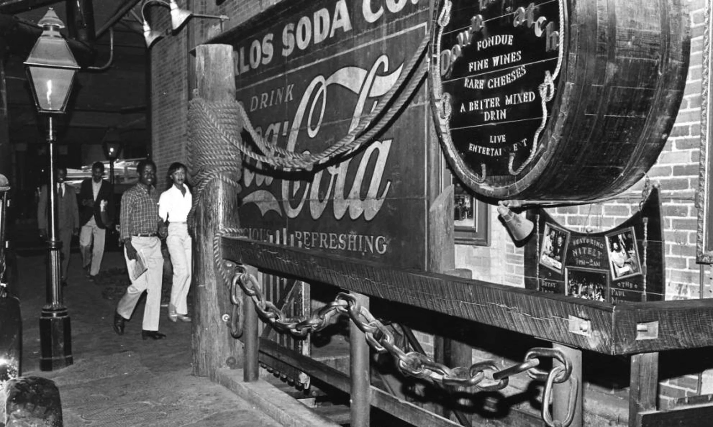 Black and white photo of a city street scene at night with three men walking past a large billboard advertising drinks and snacks. The billboard has signs with words like 'Los Soda Co.' and 'Drink Coca-Cola,' and features decorative elements like ropes and chains. A street lamp is visible to the left, and there are brick buildings in the background.
