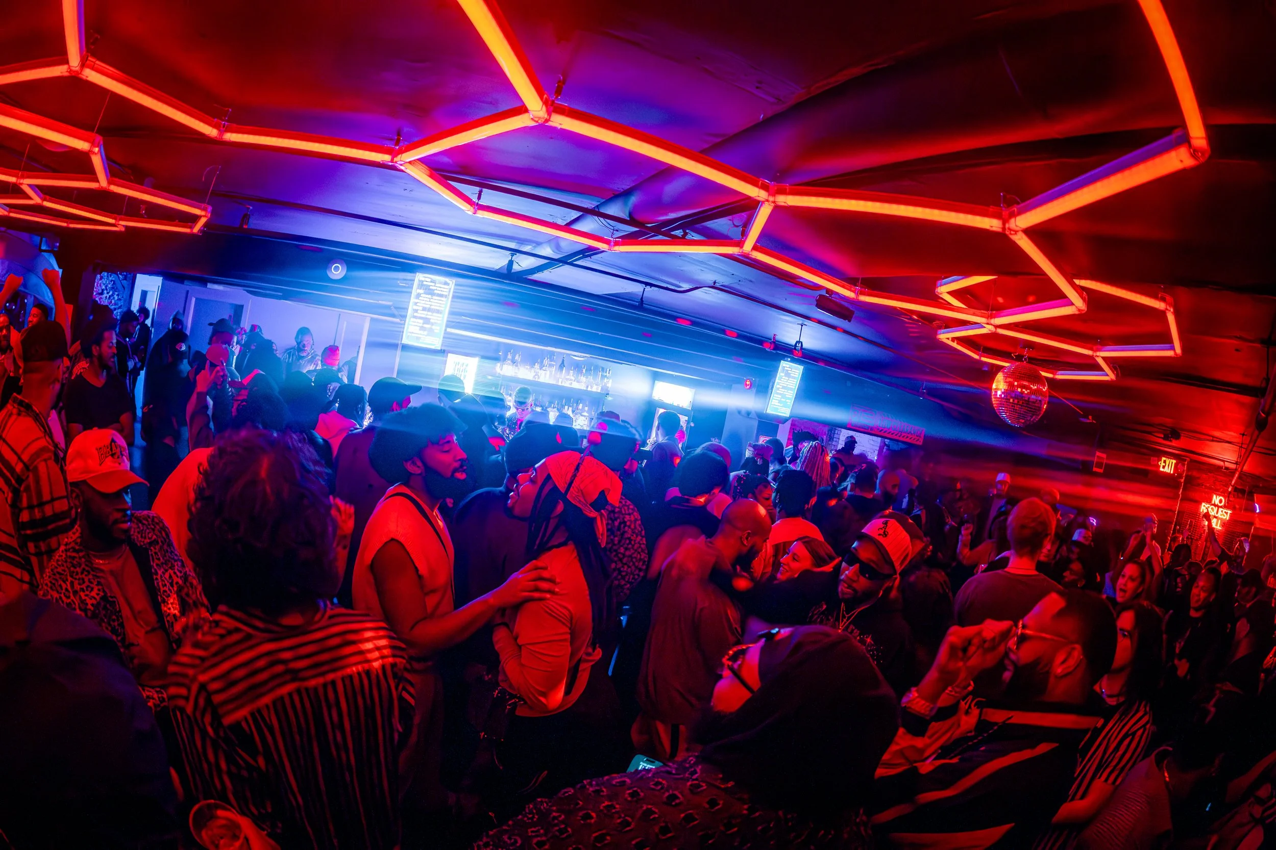 A crowded nightclub with people dancing and socializing under vibrant neon red and blue lighting, with a digital bar menu display in the background and a disco ball hanging from the ceiling.