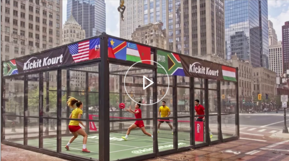 People playing kicknet in a glass-enclosed court on a city street, with tall buildings in the background.