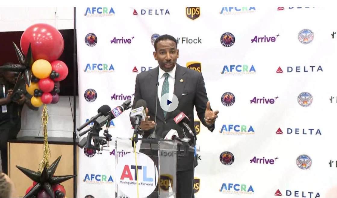 Man in a suit speaking at a press conference with microphones, with a backdrop featuring logos of AFCFA, Delta, UPS, Arrive, and ATL. Decorations include balloons and star-shaped balloons.