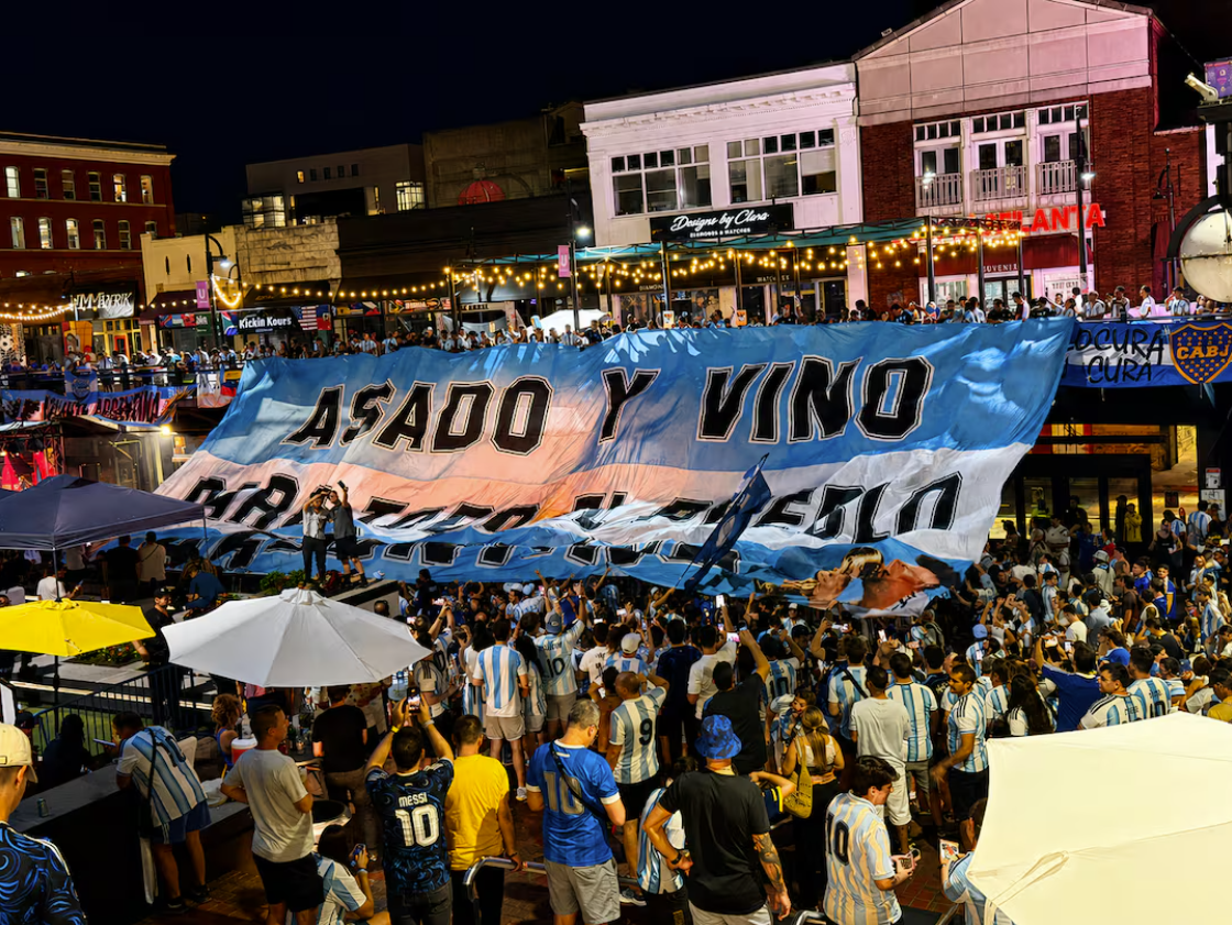 Crowd of people gathered at an outdoor event holding a large banner that reads "Asado y vino" in a lively urban setting at night.