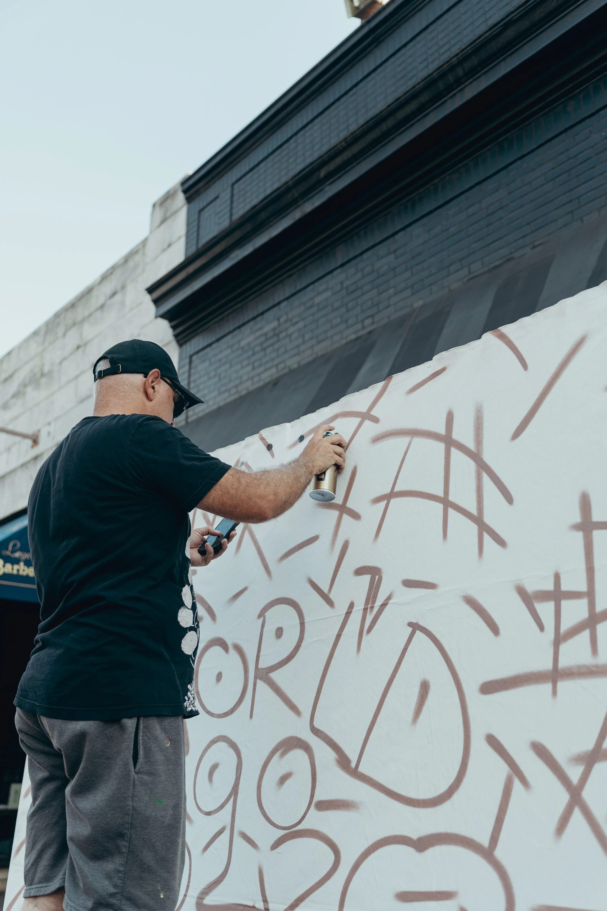 Man wearing a black cap, black T-shirt, and gray shorts appears to be spray painting on a large white canvas or wall with abstract brown lines and shapes outdoors under a blue sky.