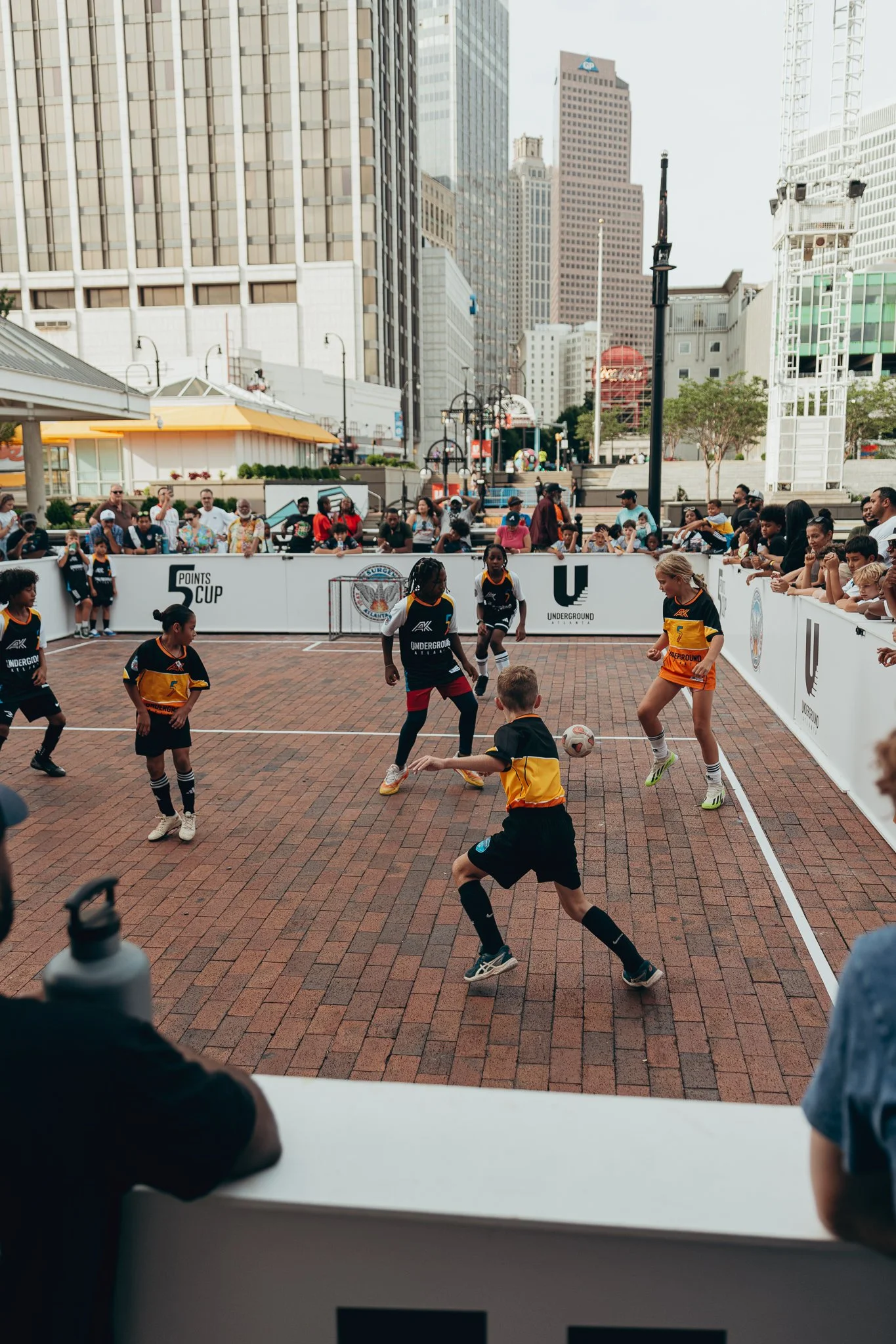 Children playing indoor soccer on a brick court surrounded by a white barrier with logos, with spectators watching behind the barrier, and a cityscape with tall buildings in the background in Downtown Atlanta - Underground Atlanta.