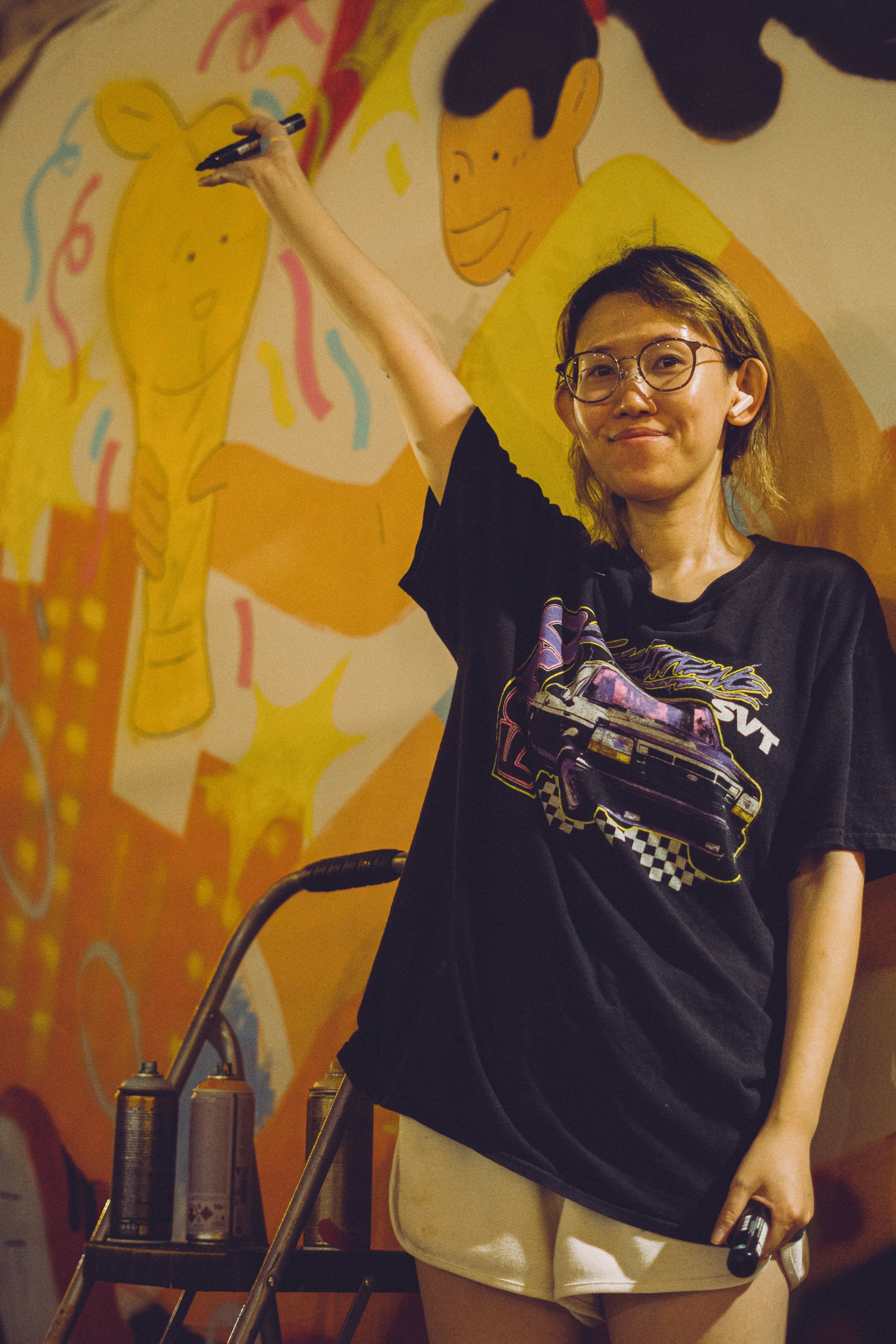 A person with short hair and glasses wearing a black t-shirt with a car graphic and beige shorts, holding a marker and standing in front of a colorful mural with cartoon faces of a girl and a boy. Part of a cart with spray cans is visible in the foreground.