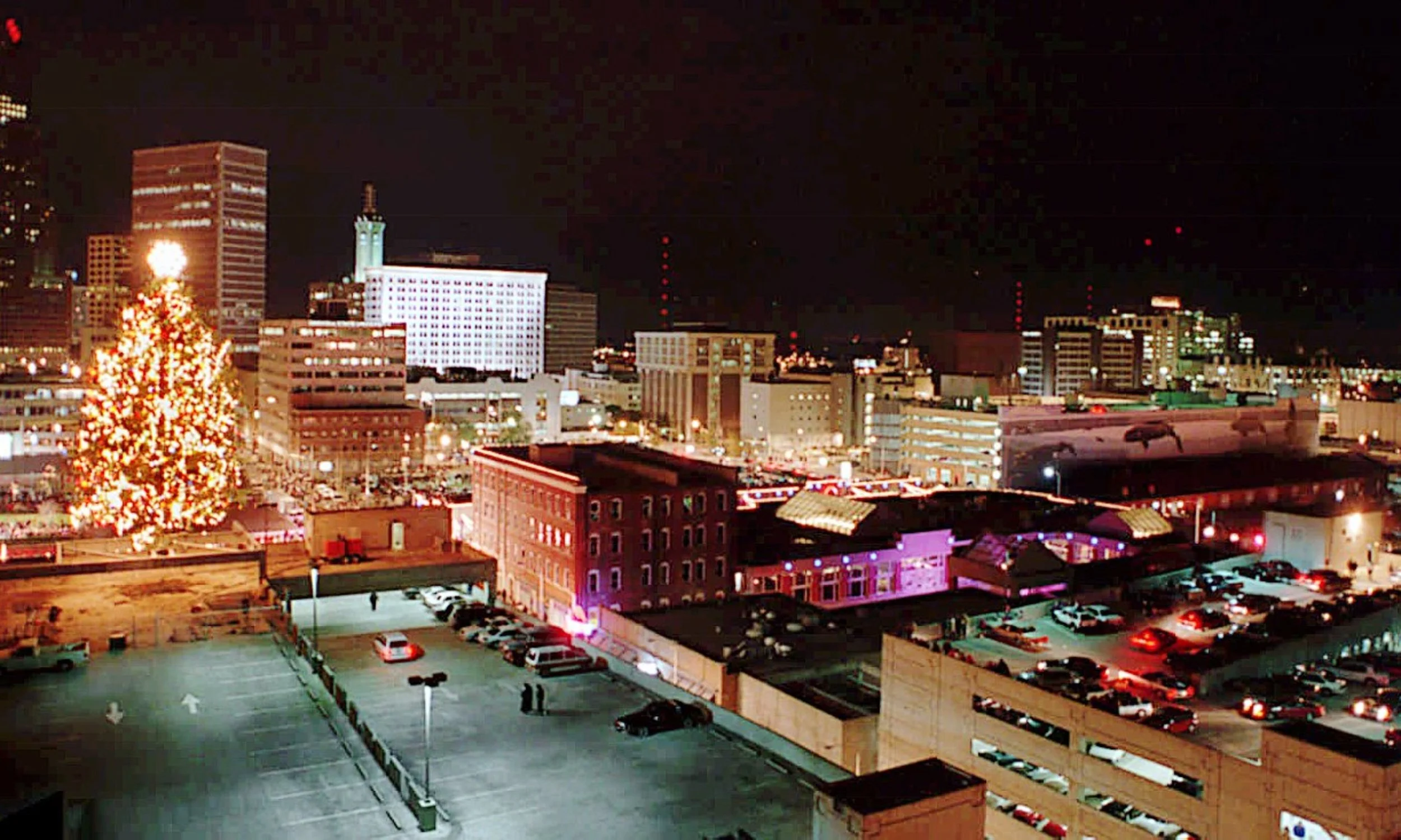Nighttime cityscape with illuminated buildings, a large decorated Christmas tree in the foreground, and a parking lot with cars.