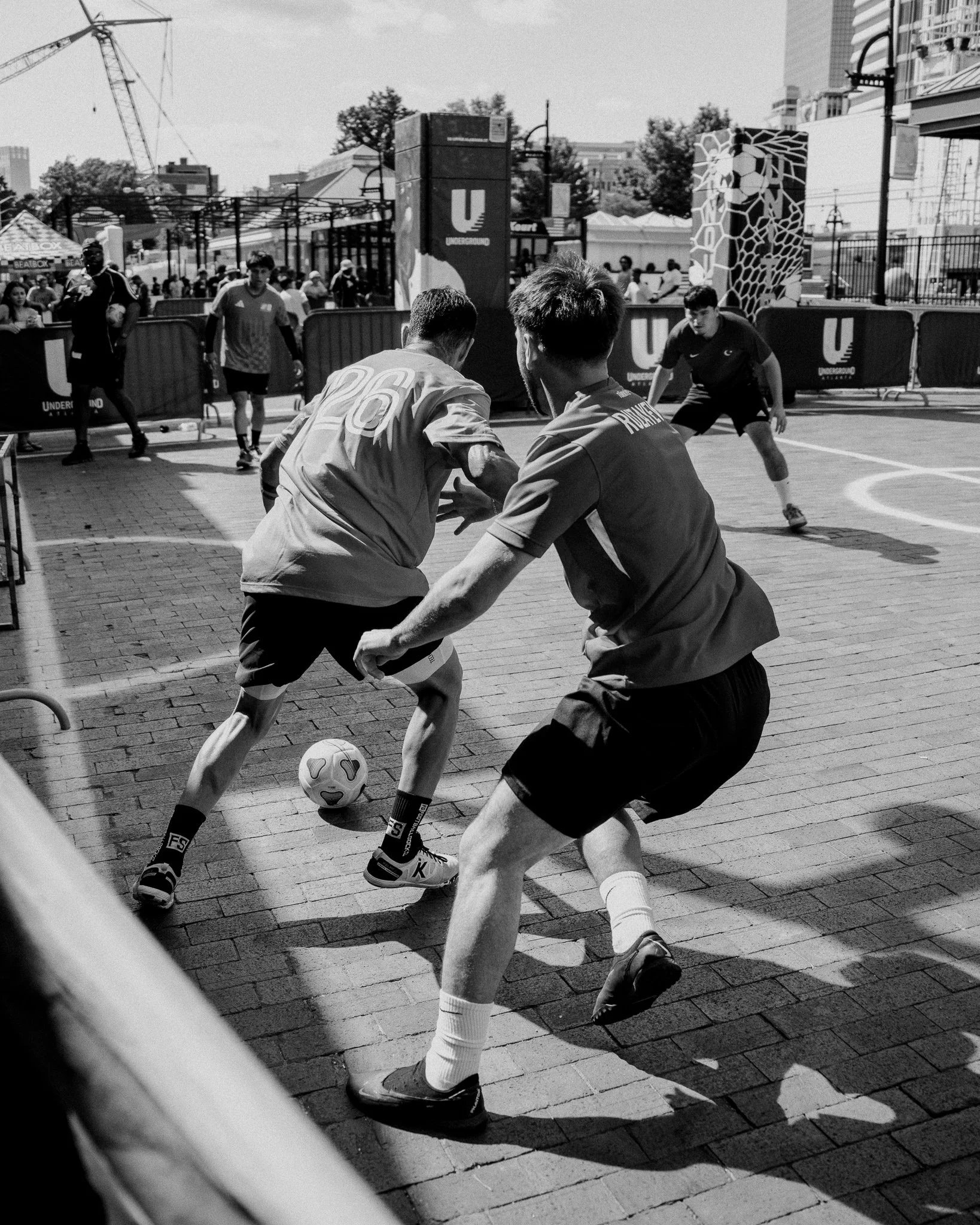 A black-and-white photo of several young men playing outdoor soccer on a city street, with some spectators watching behind a barricade. The players are in motion, competing for the ball, with one player in the foreground wearing a jersey with the number 26. City buildings and a crane can be seen in the background.