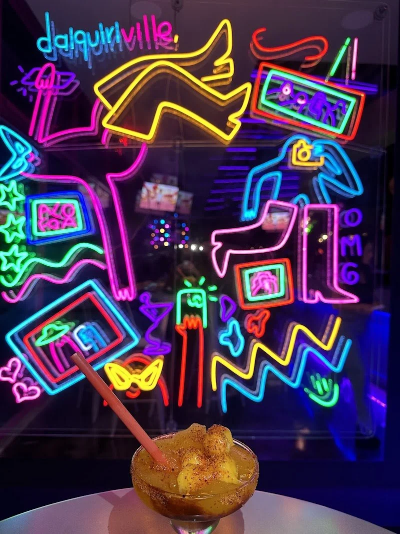 Colorful neon sign with various abstract shapes, figures, and words, including 'daiquiri villa,' with a drink in the foreground topped with fruit and a straw.