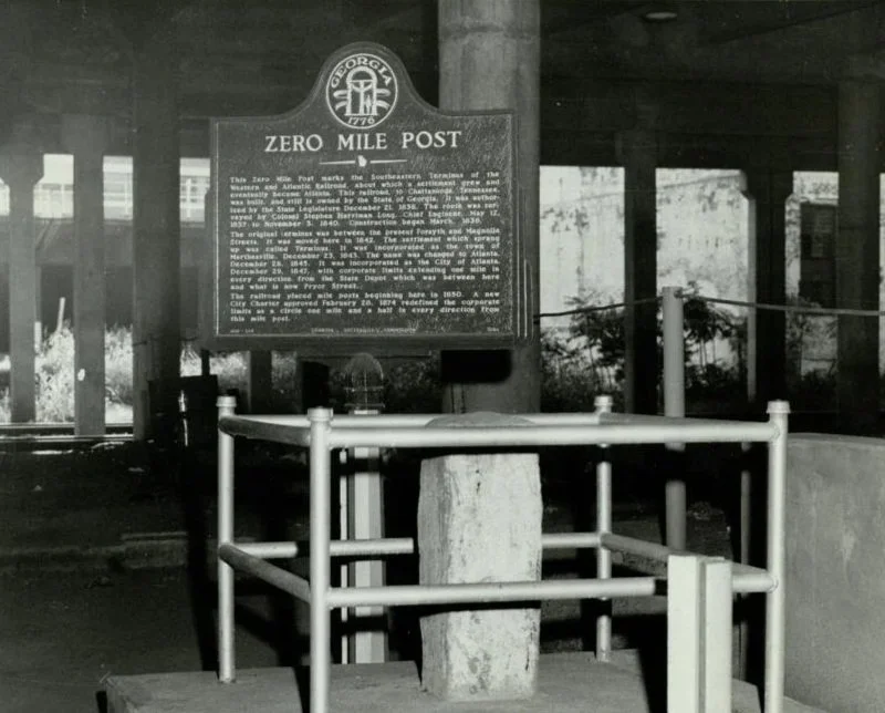 Historical marker sign at the Zero Mile Post, with informational text and a nearby railing, under a bridge or overpass.