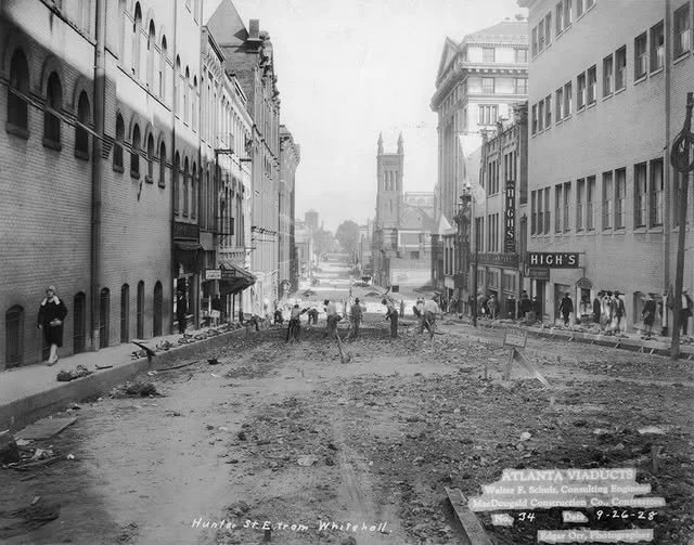 Black and white photo of a city street under construction, with people walking along the sidewalks and on the uneven, dirt road. Buildings line both sides, and a bridge is visible in the distance.