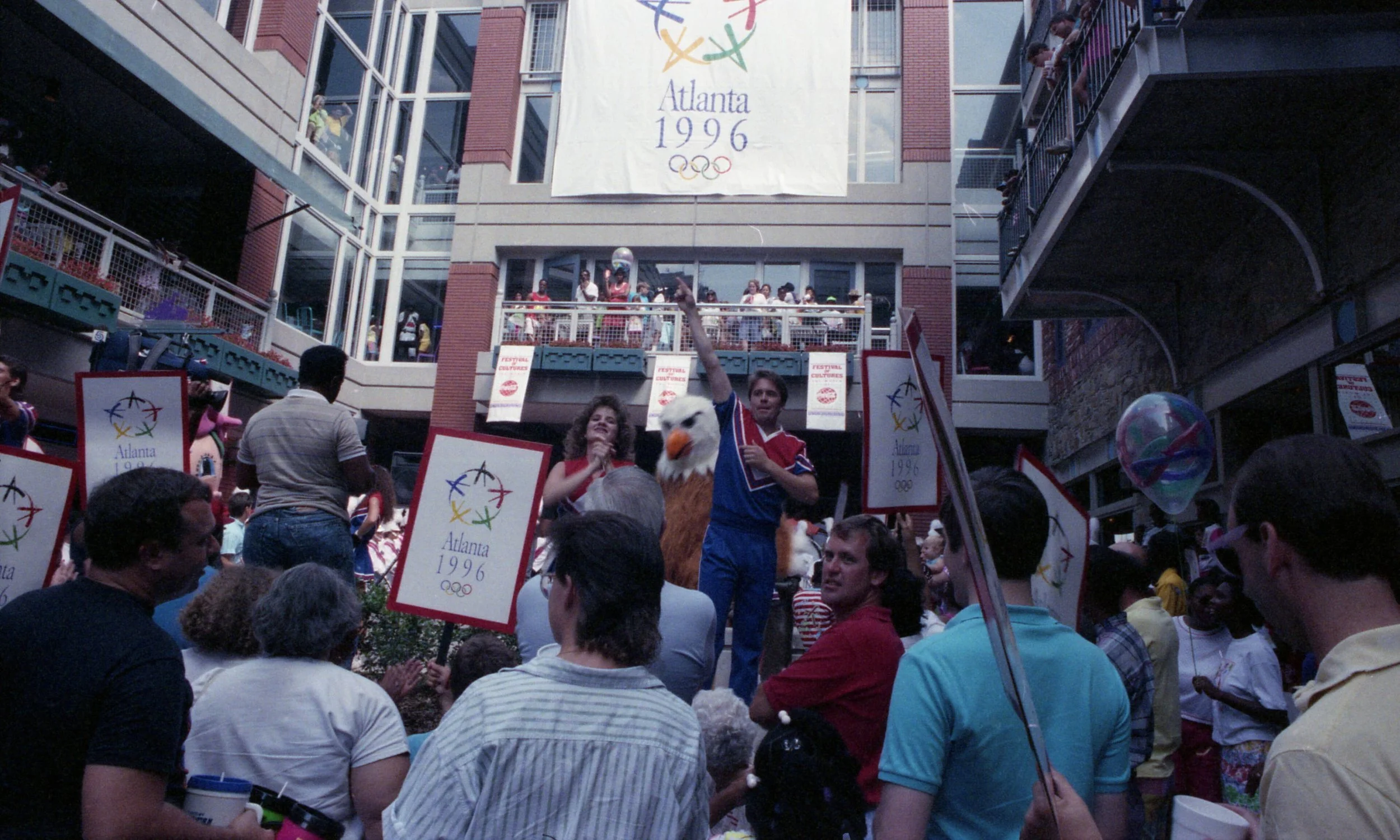 A crowd gathers at an outdoor event in Atlanta during the 1996 Summer Olympics. People hold flags and banners with the Olympic rings and the words 'Atlanta 1996,' with a large Olympic flag hanging above. A person dressed as an eagle mascot is visible among the crowd.