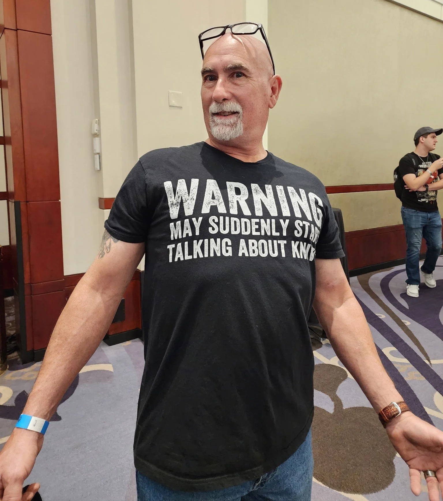 A man with a goatee and glasses on his head, wearing a black T-shirt with a humorous warning message, stands in a conference room. There are two people in the background, one wearing a black graphic T-shirt and cap, holding a smartphone.