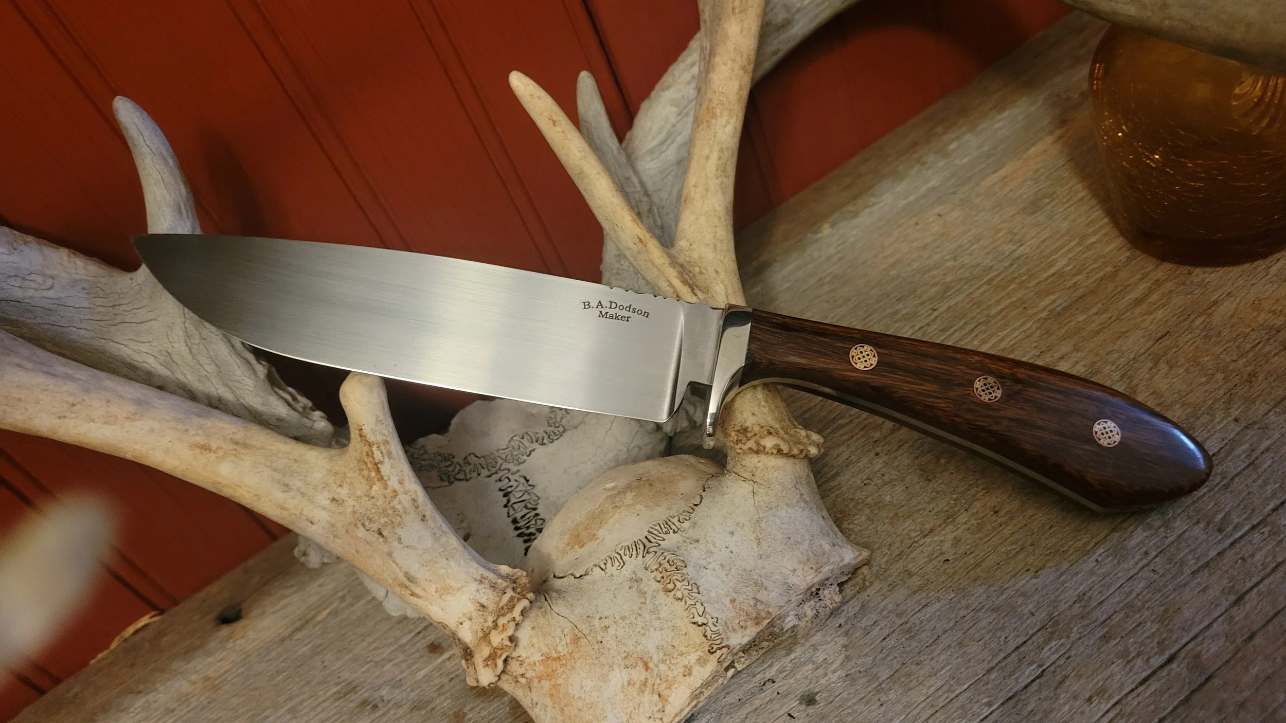 A kitchen knife with a stainless steel blade and a wooden handle is resting on an antler with a deer skull.
