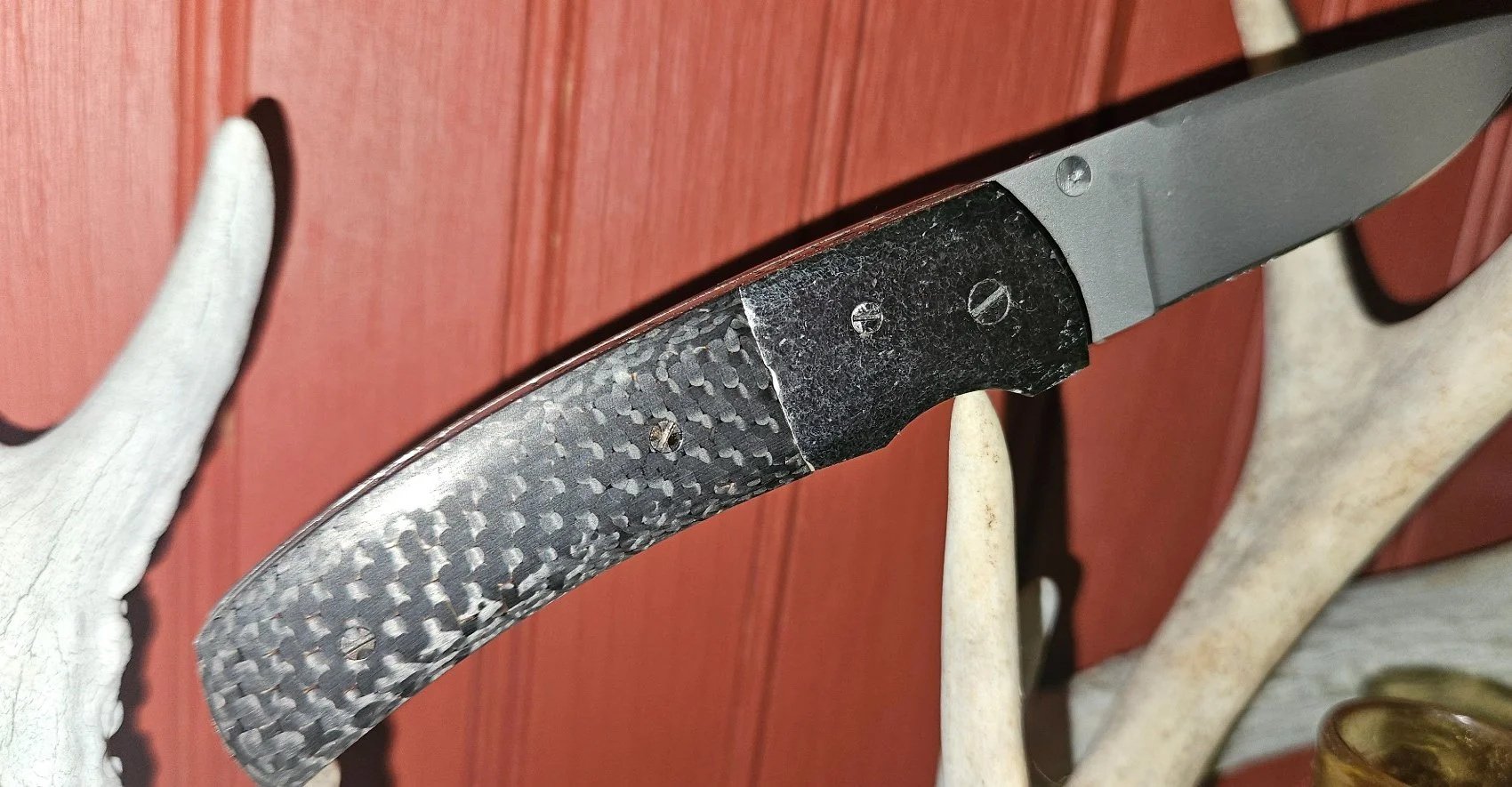 Opposite-side view of a B.A. Dodson carbon fiber and tooled titanium liner lock folder displayed on antlers, showing the carbon fiber handle and textured titanium bolster.