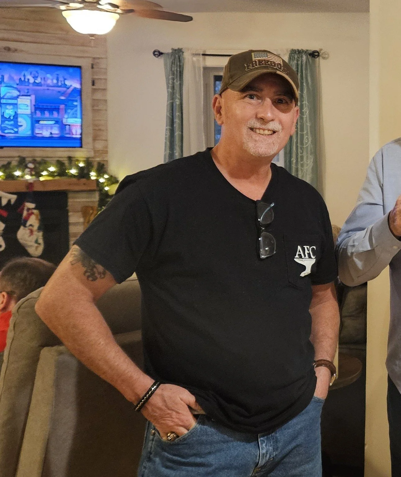 A smiling man with a beard and mustache wearing a tan cap with an American flag patch, a black t-shirt with an alcohol-related logo, and jeans, standing indoors in a living room with a fireplace, stockings, and holiday decorations in the background.